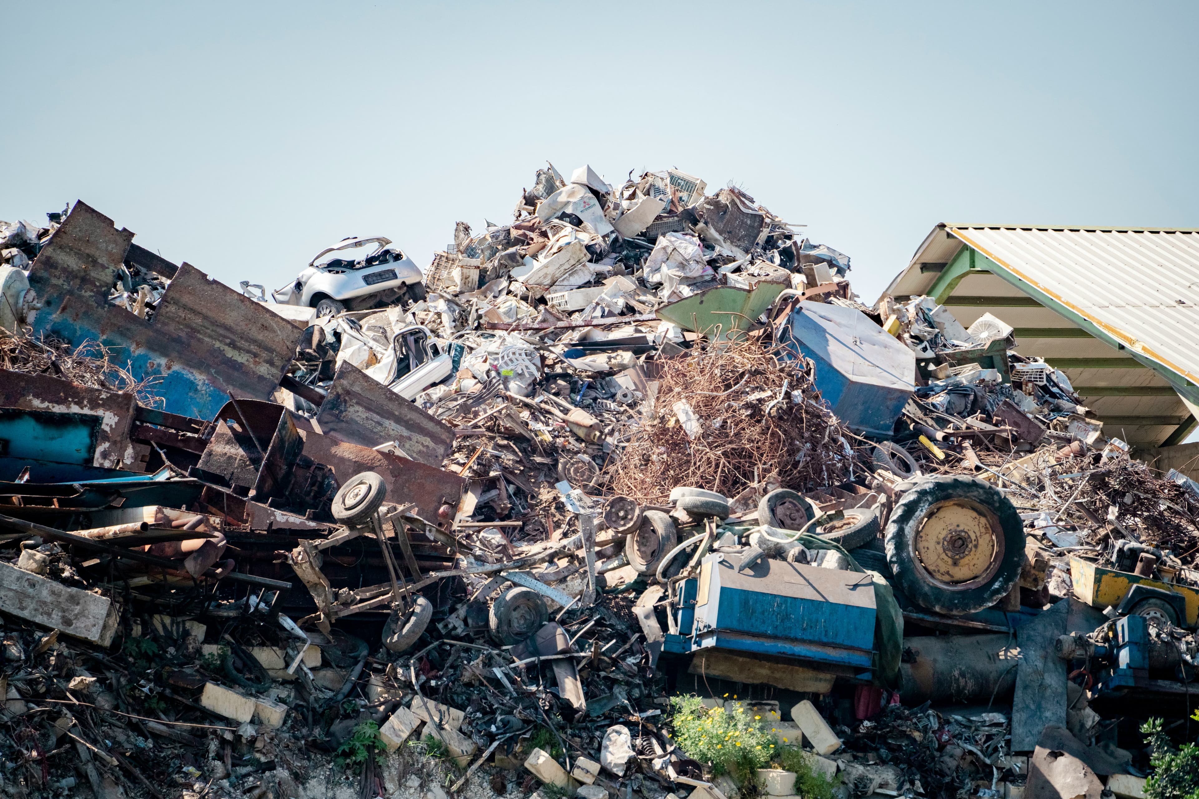 A tall heap of mixed scrap metal, appliances and vehicle parts in a scrapyard under a clear sky