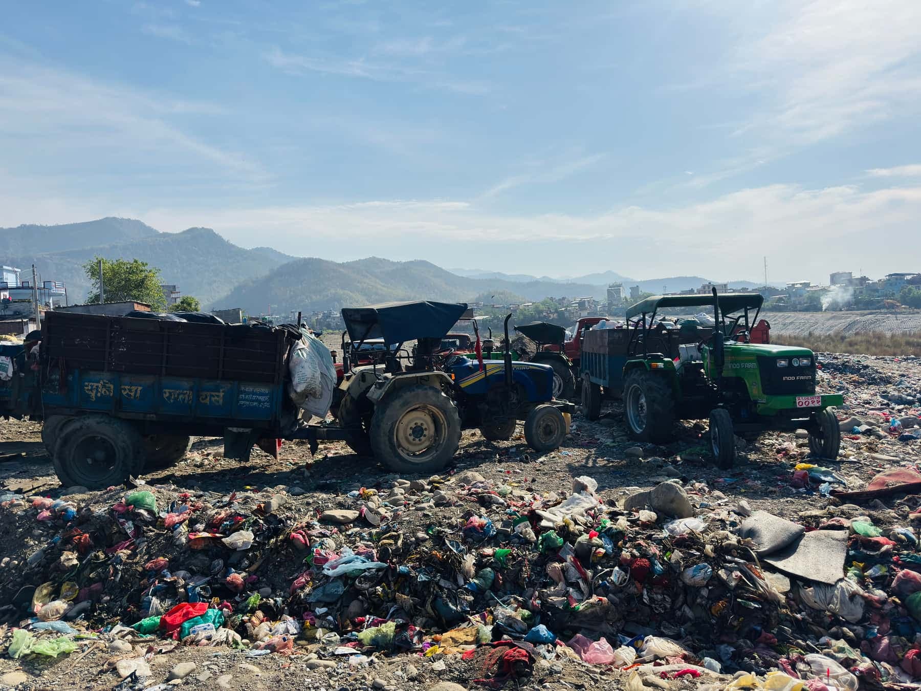 Two tractors with trailers parked on a waste ground strewn with mixed plastic, with hills in the background