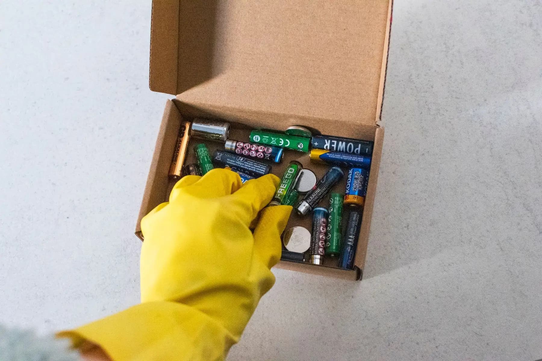 A gloved hand placing used batteries into a cardboard collection box